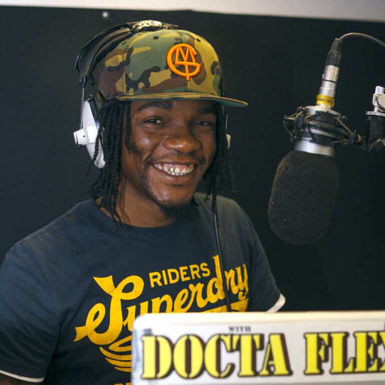 a man in a cap and headphones stands smiling in front of a microphone